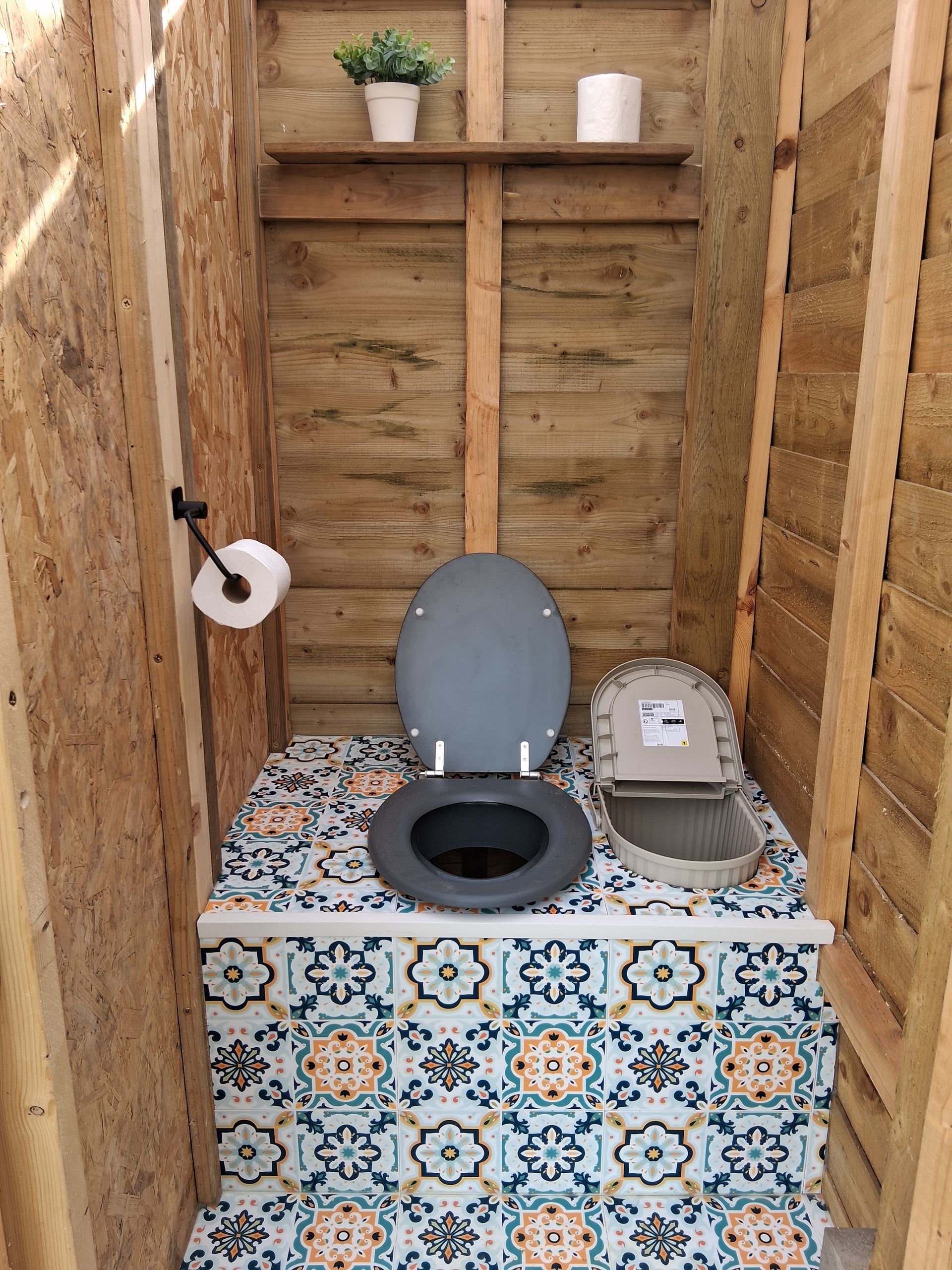 Interior of an outhouse with a toilet, shelf with plants and toilet paper, walls are wood.