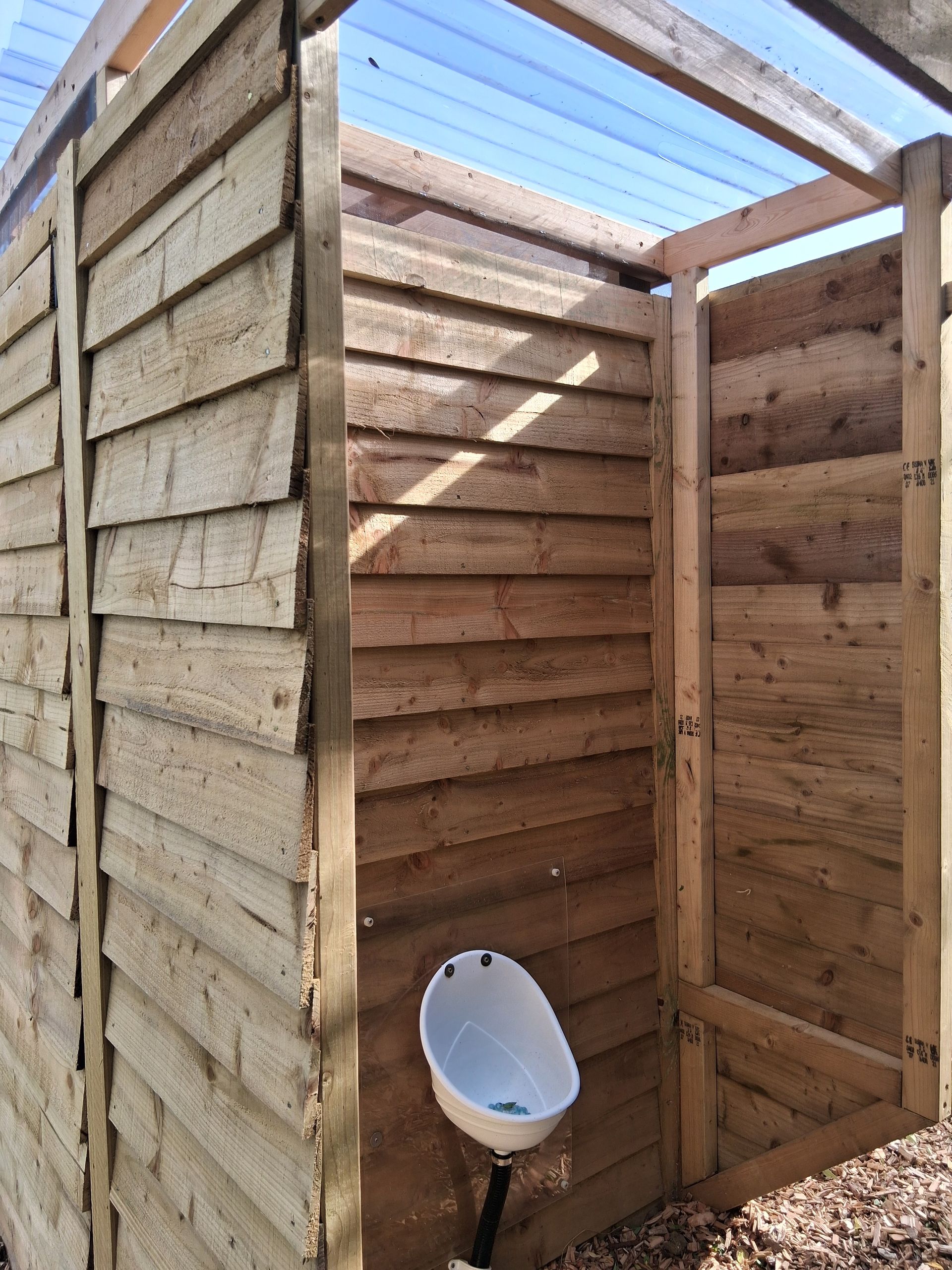Outdoor wooden urinal stall. Wood planks, white urinal, brown frame, blue sky.