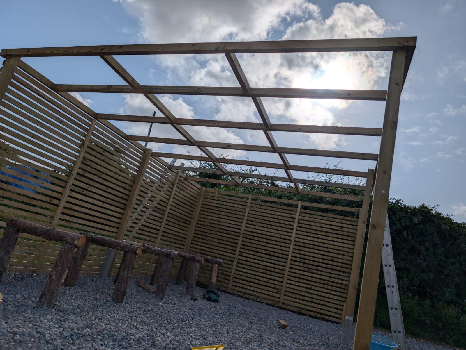 Wooden pergola frame with slatted fence panels, built outdoors under a partly cloudy sky.