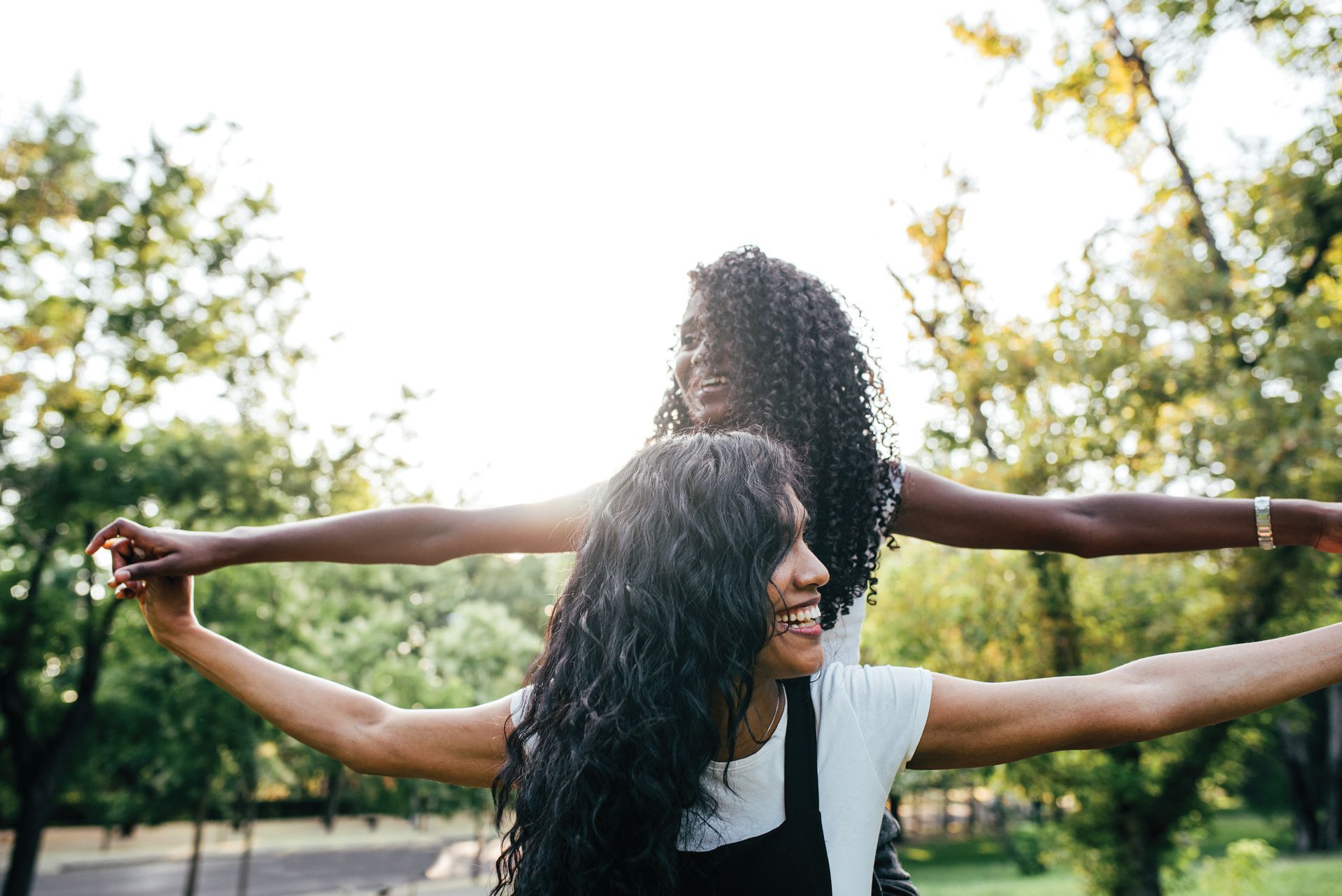 Two women are standing next to each other in a park with their arms outstretched.