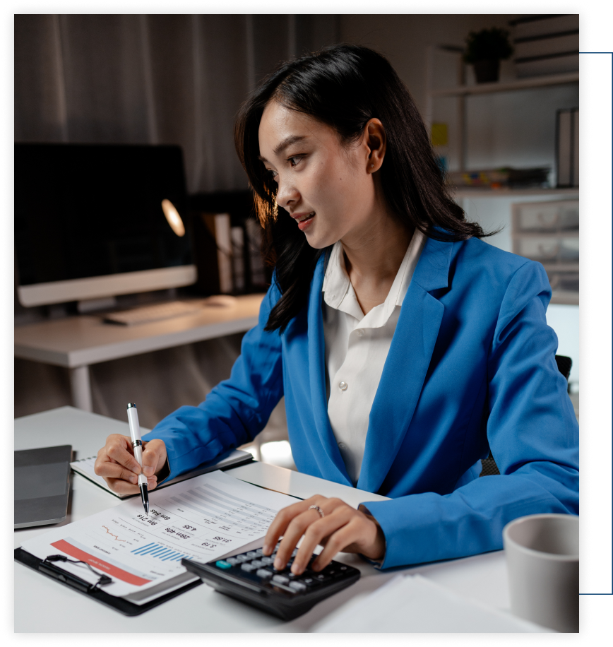 A woman in a blue jacket is sitting at a desk using a calculator.