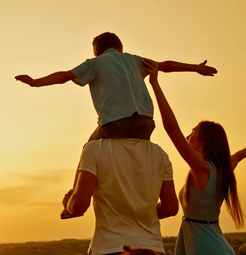 Two people carry a child on their shoulders at sunset, arms outstretched in a field.