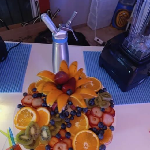 Una bandeja con rodajas de naranja, fresas, kiwis y arándanos, con un dispensador de nata montada al fondo.