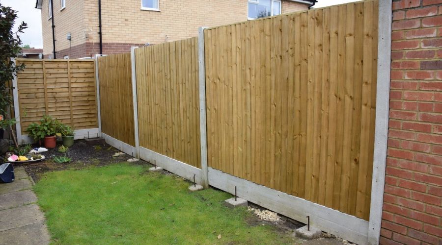 A newly installed wooden garden fence with concrete posts and gravel boards bordering a lawn next to a brick wall.