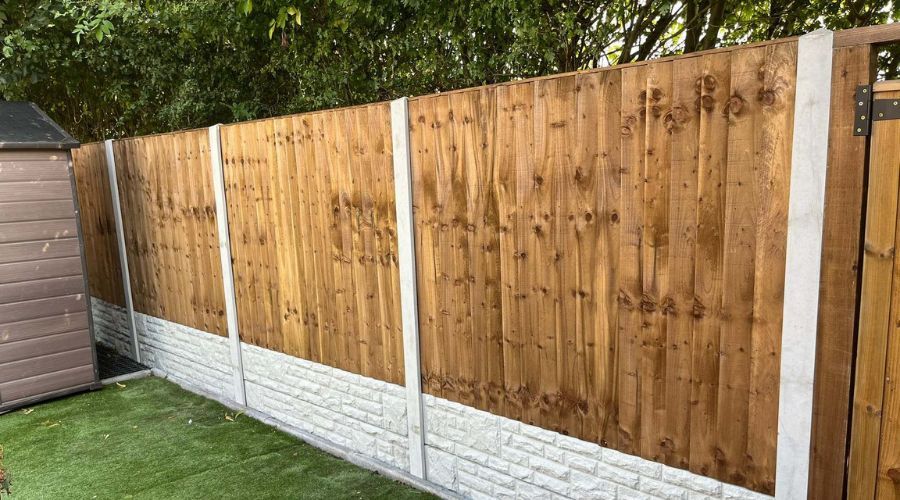 A new wooden panel fence with concrete posts and decorative rock-face gravel boards installed along an artificial grass lawn.