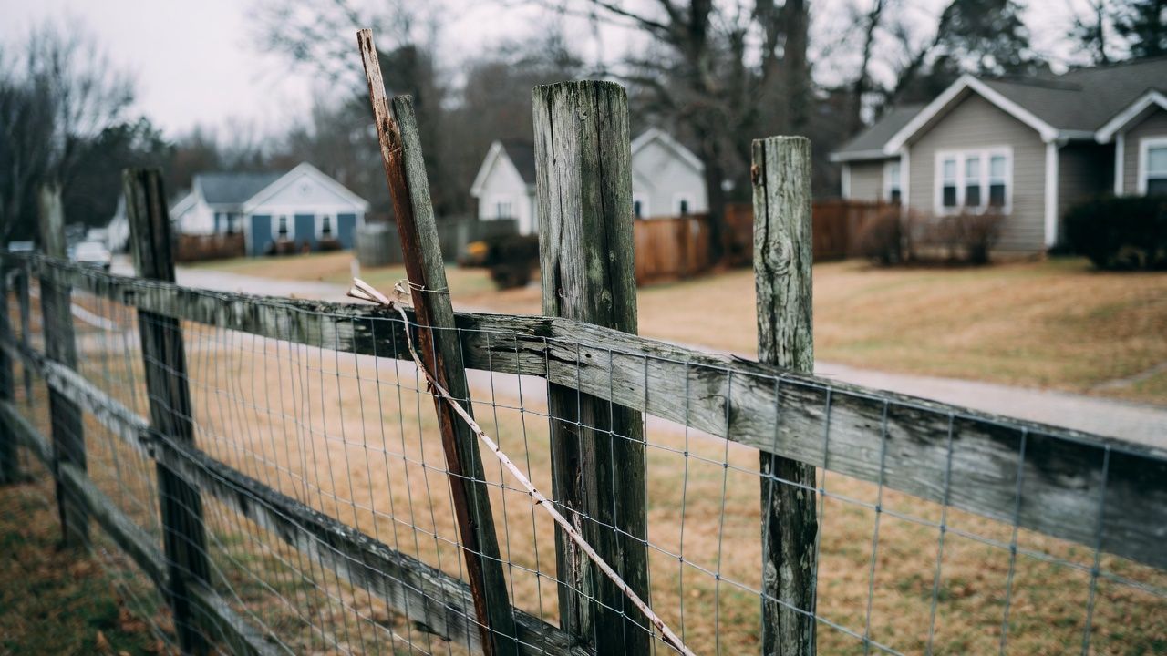 Weathered wooden fence with wire mesh in foreground; houses and road in the background.