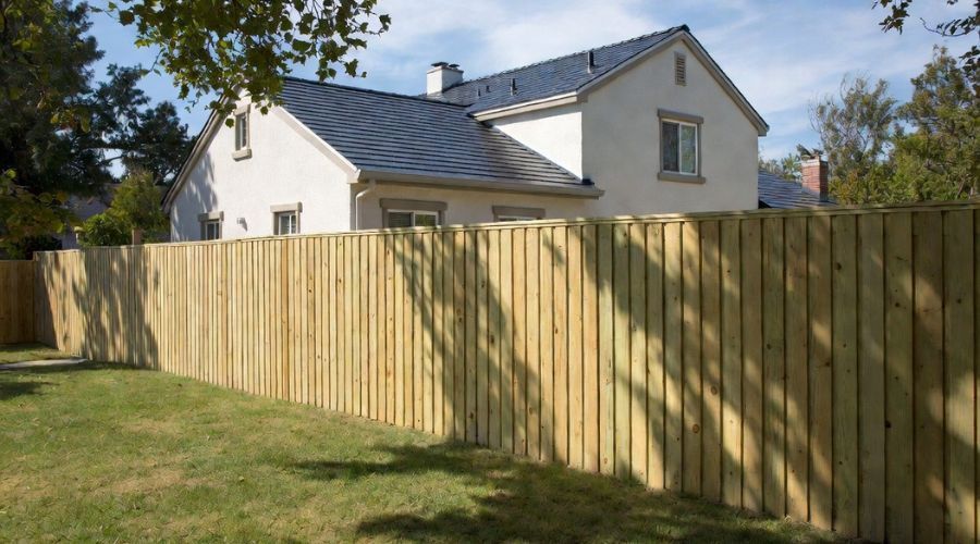 A light-colored house with a dark roof stands behind a tall, light wood picket fence in a grassy yard.