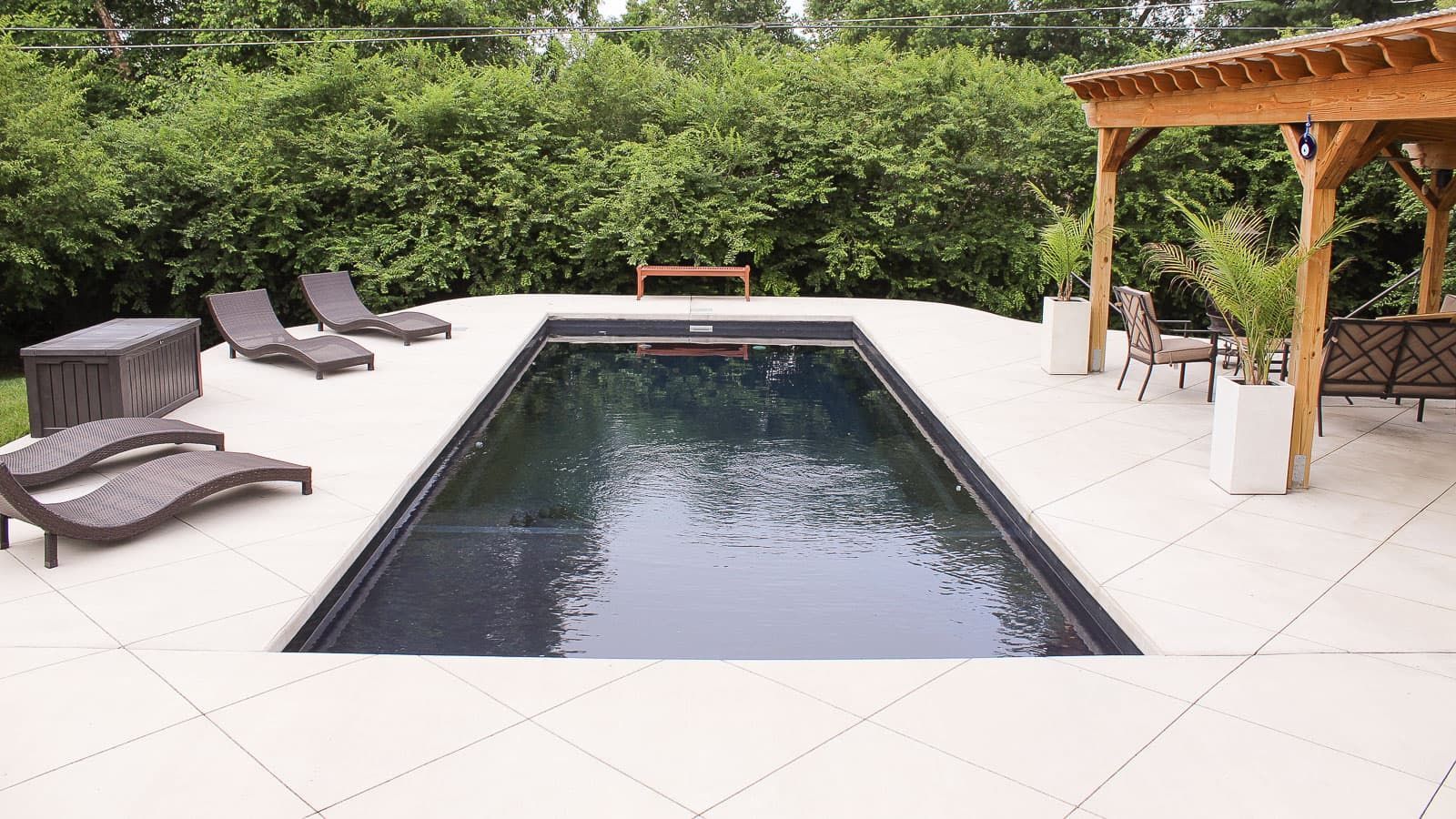 A large swimming pool in the Freedom + Splash Pad Layout in Volcanic Black surrounded by chairs and a pergola. 