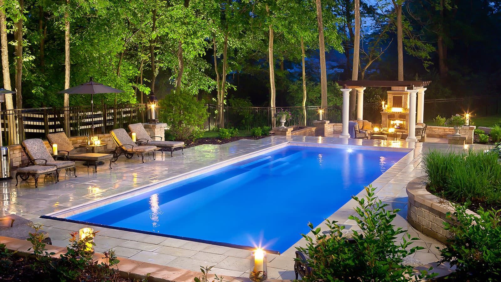 A large swimming pool is lit up at night in a backyard surrounded by trees.