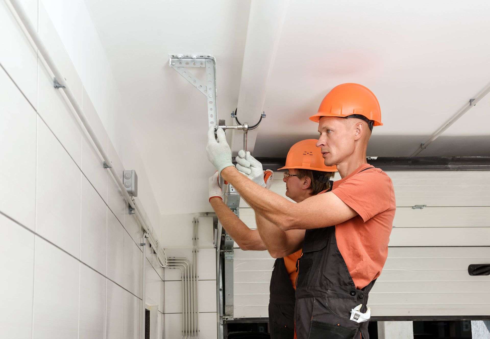 Two workers in orange hard hats install a bracket on a white ceiling.