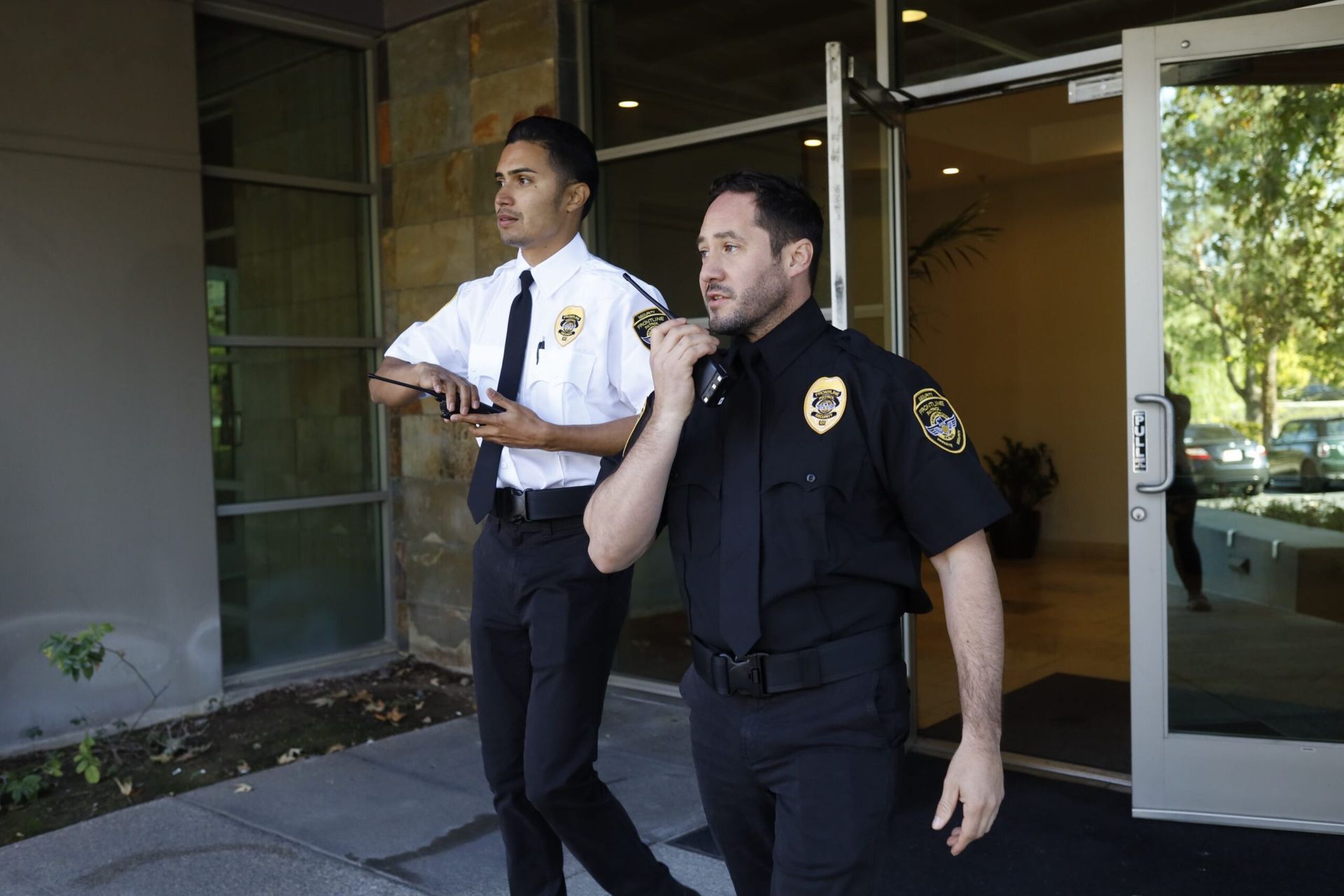 Two security guards outside a building. One looks ahead, holding a phone. The other speaks into a radio.