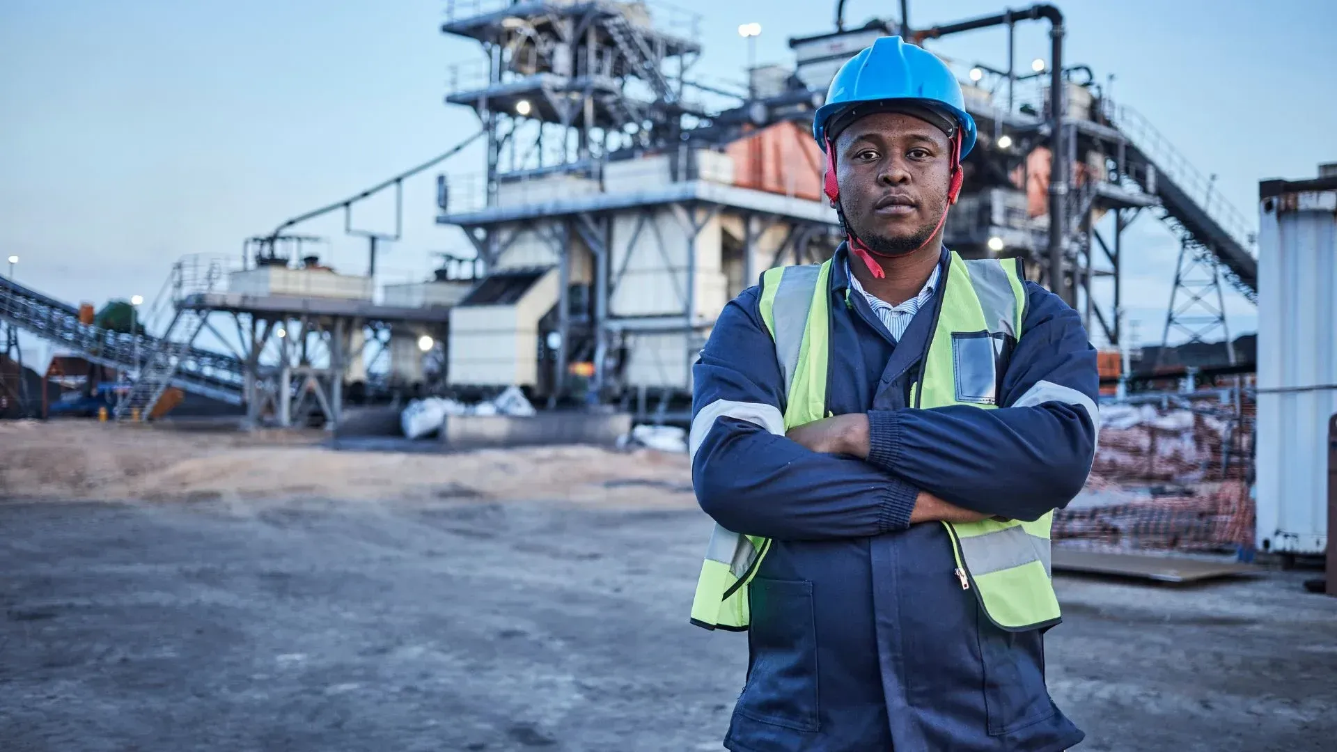 Construction worker in safety gear stands in front of a factory, arms crossed, looking at the camera.