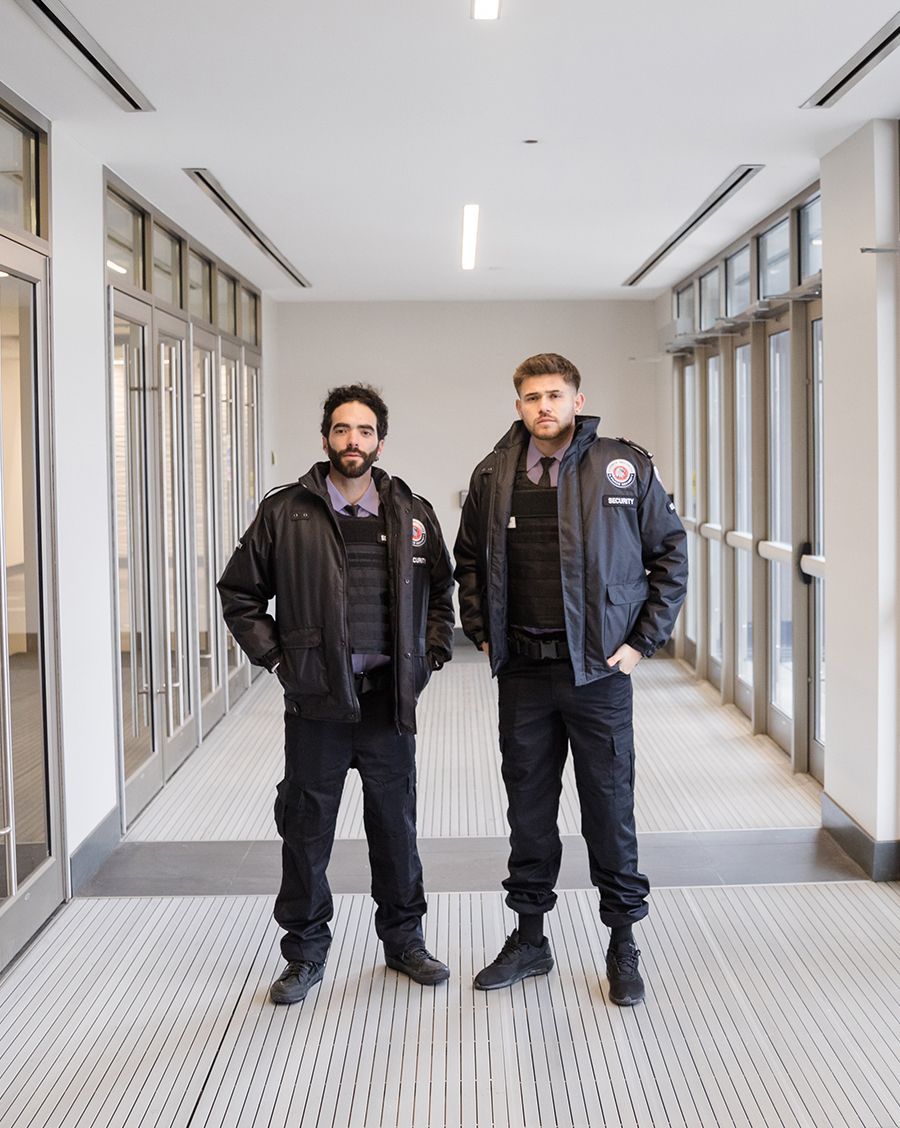 Two security guards in uniform stand in a hallway, hands on hips. They wear black jackets and vests.
