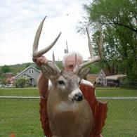 A person holds a taxidermy deer mount with large antlers, standing outdoors in front of a residential area.