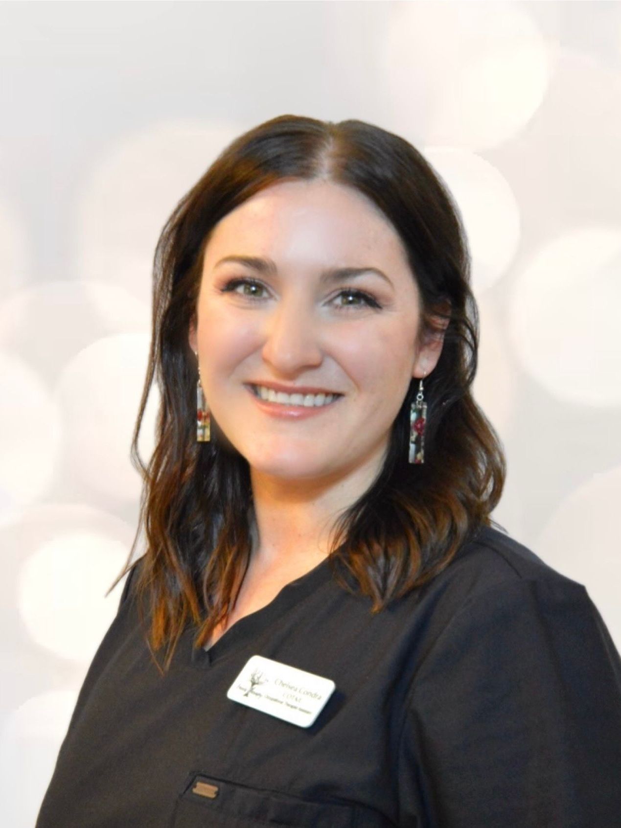 A woman wearing a black scrub top and earrings smiles for the camera.