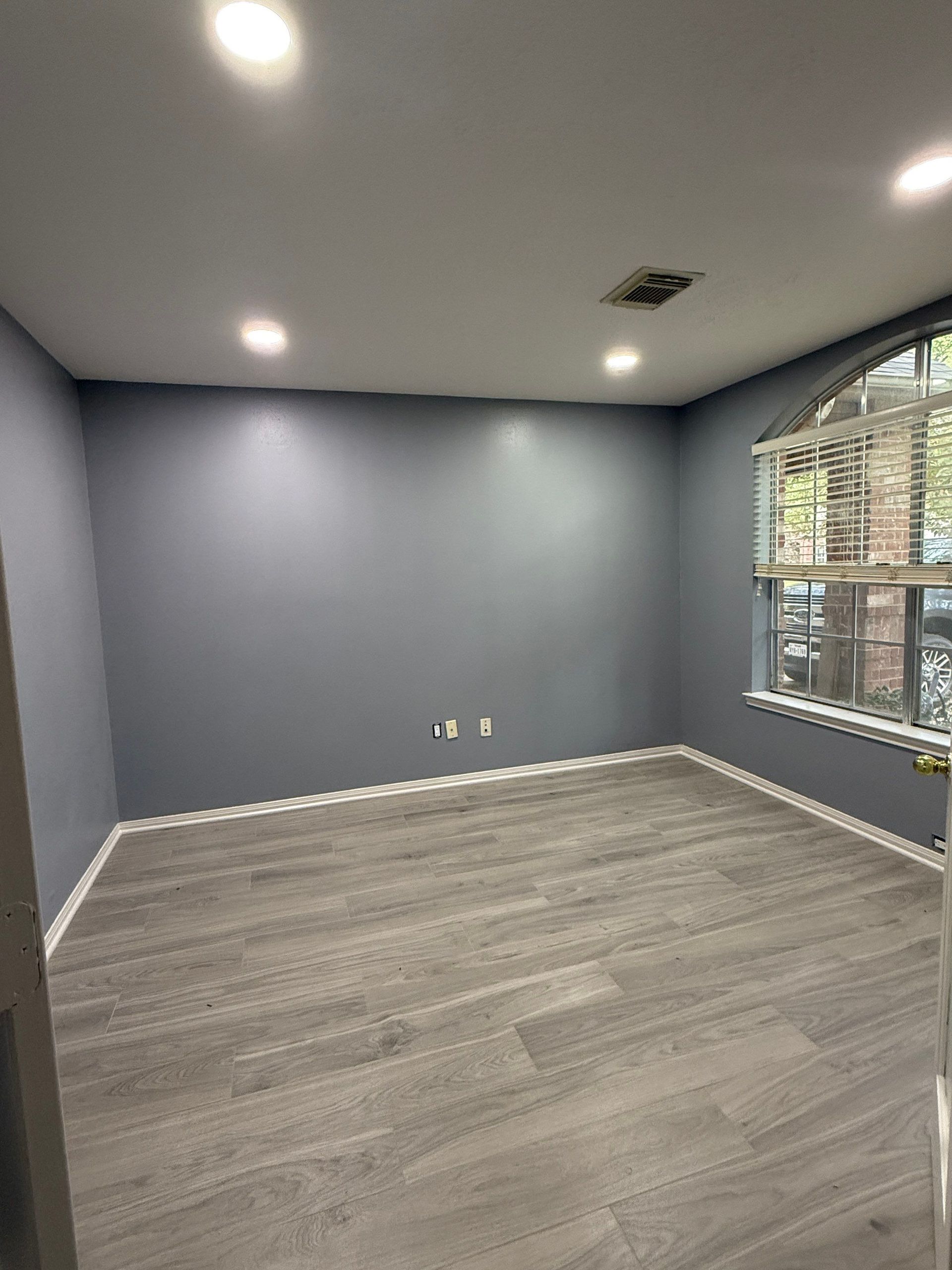Empty room with gray walls, gray wood-look floor, and a rounded window. Ceiling lights are on.