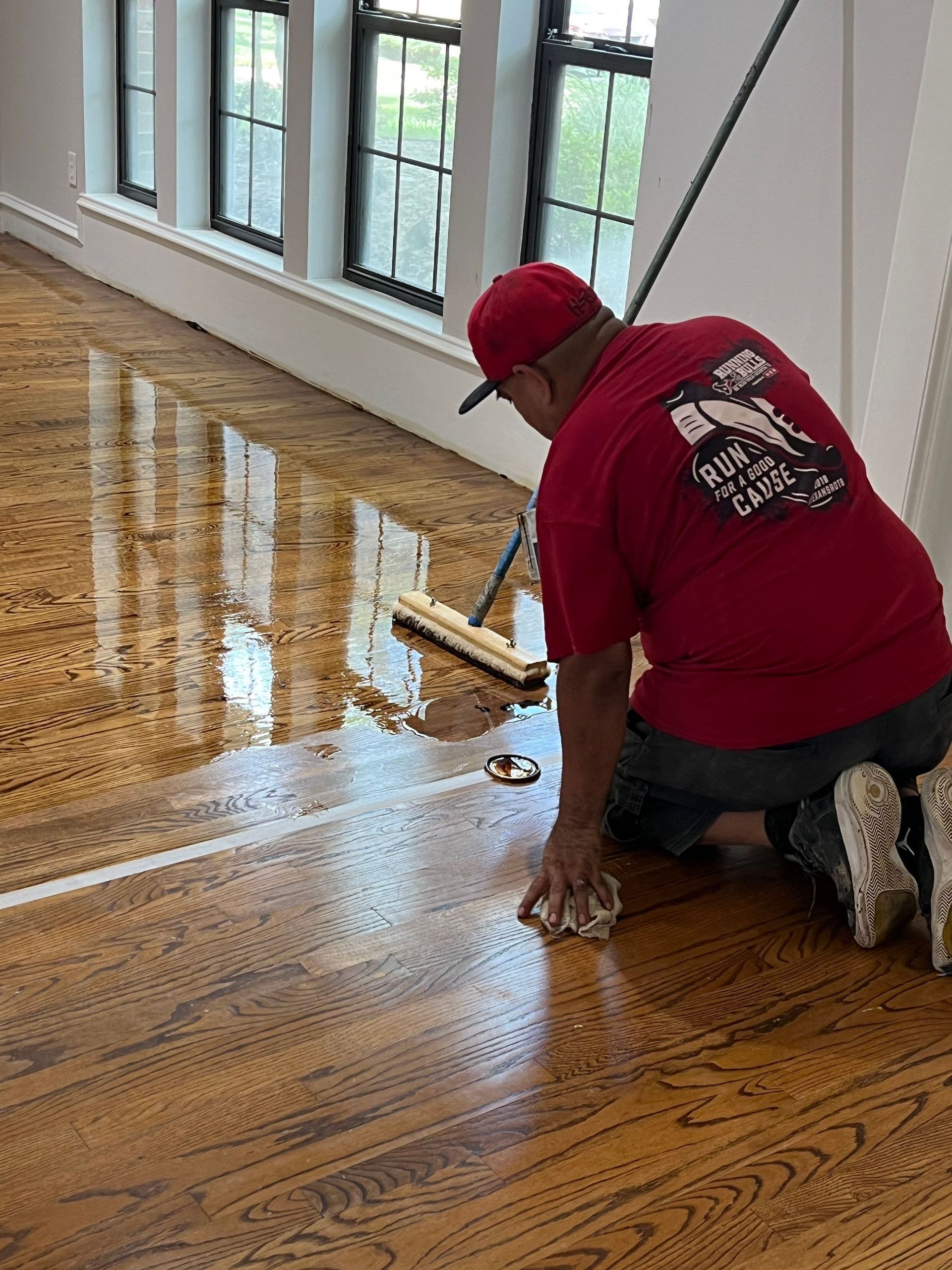 Person kneeling, applying finish to hardwood floor with mop and cloth. Interior shot, windows in background.