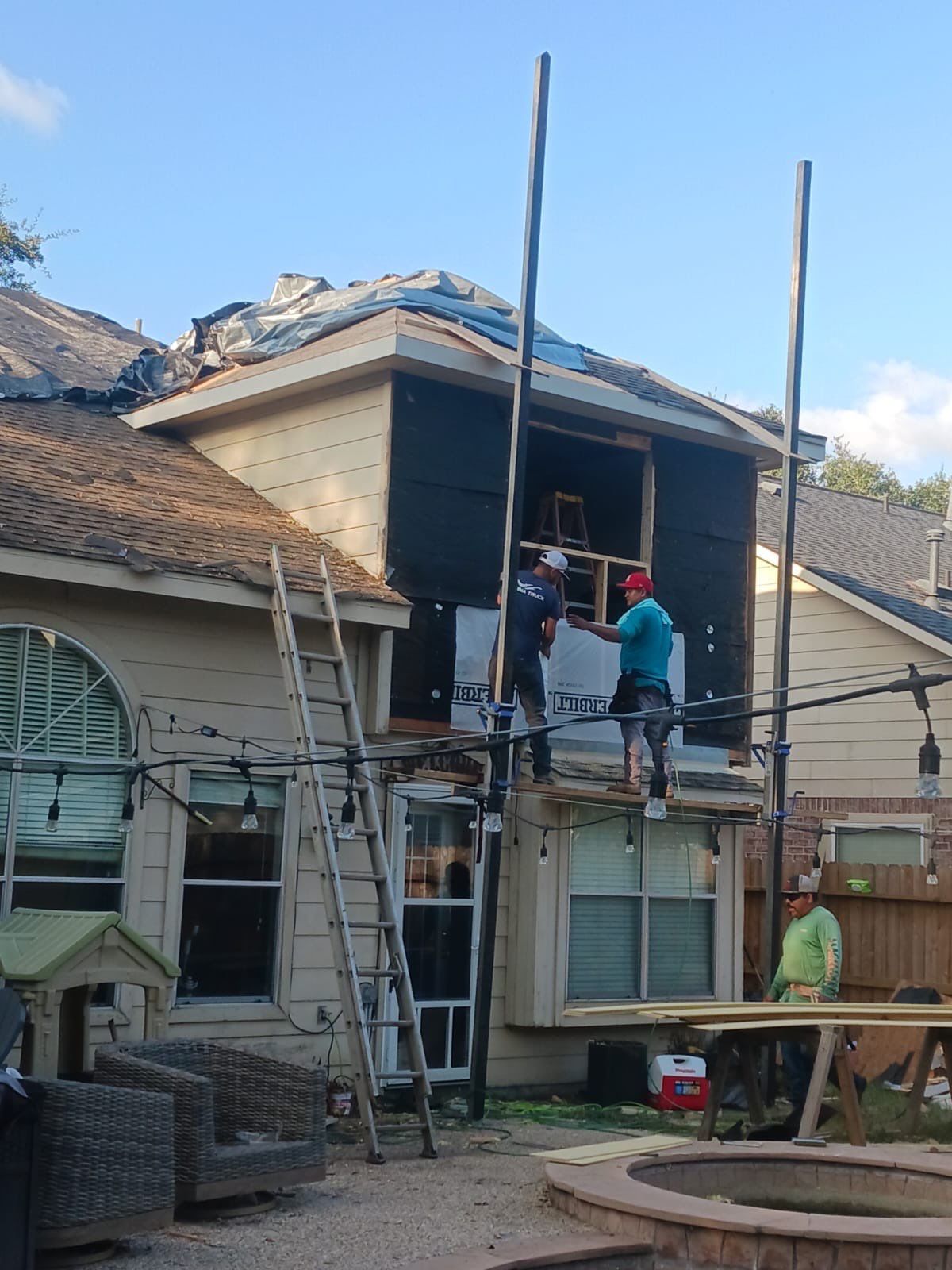 Two workers on scaffolding repair a house exterior. A ladder leans against the structure.
