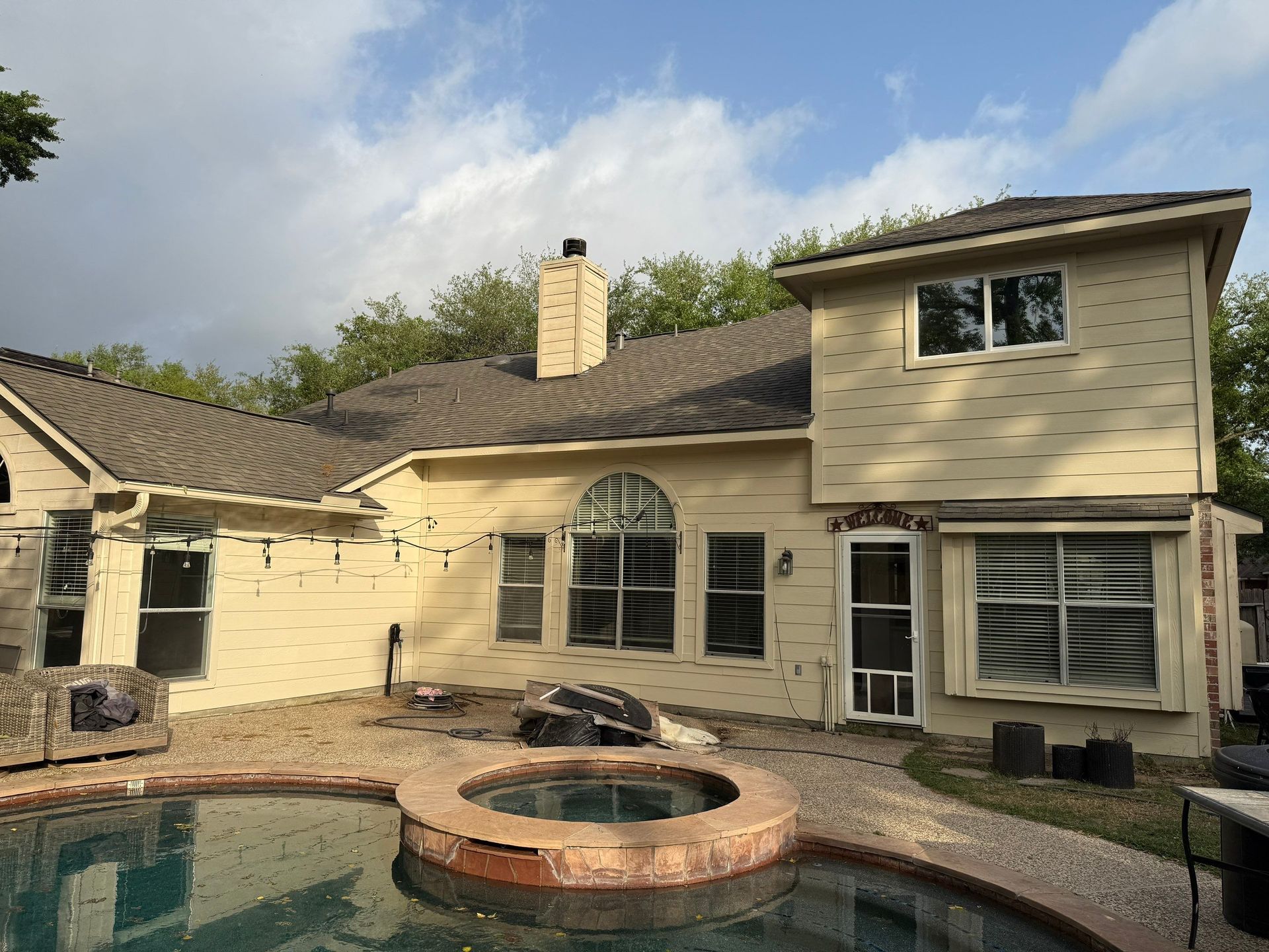 Backyard view of a two-story beige house with a pool and a hot tub. Overcast sky.