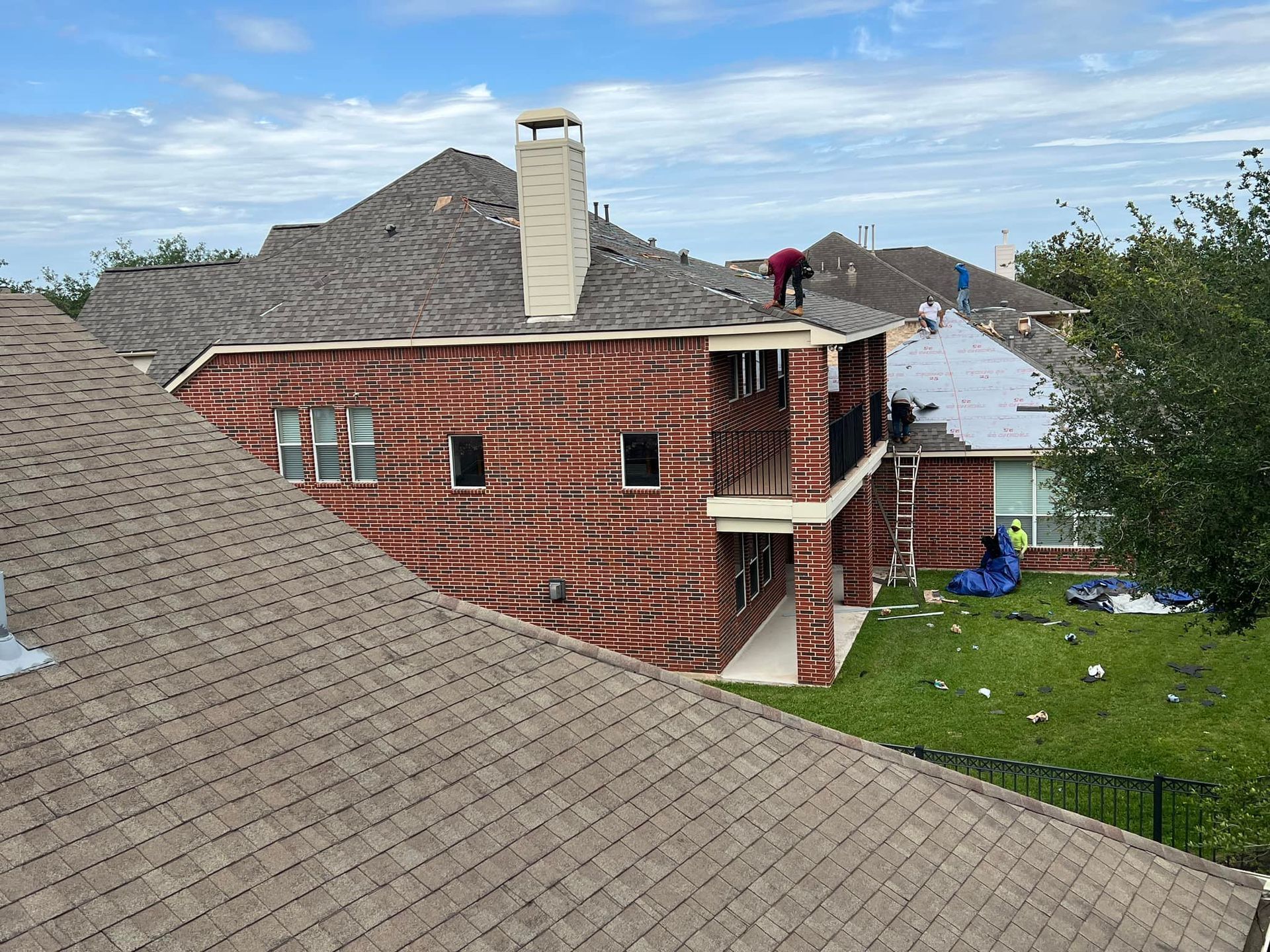Roofers working on a brick house with brown shingle roof, green grass, and blue sky.