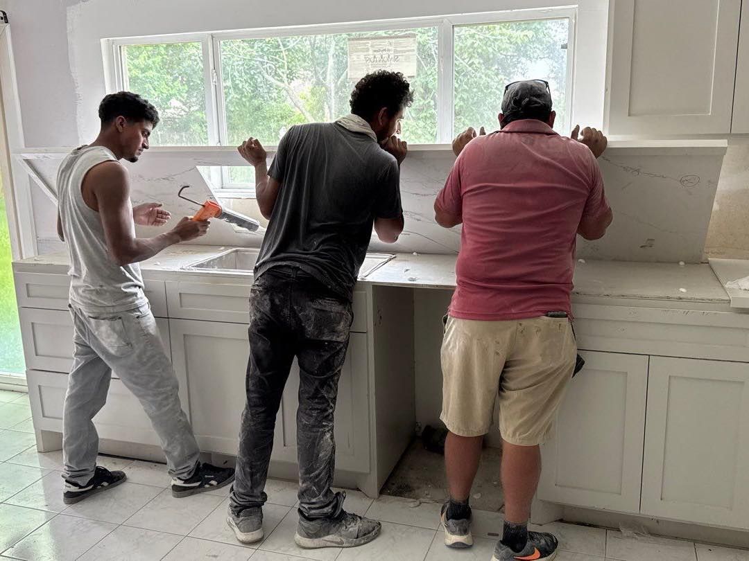 Three workers installing white countertops over white cabinets near a window.