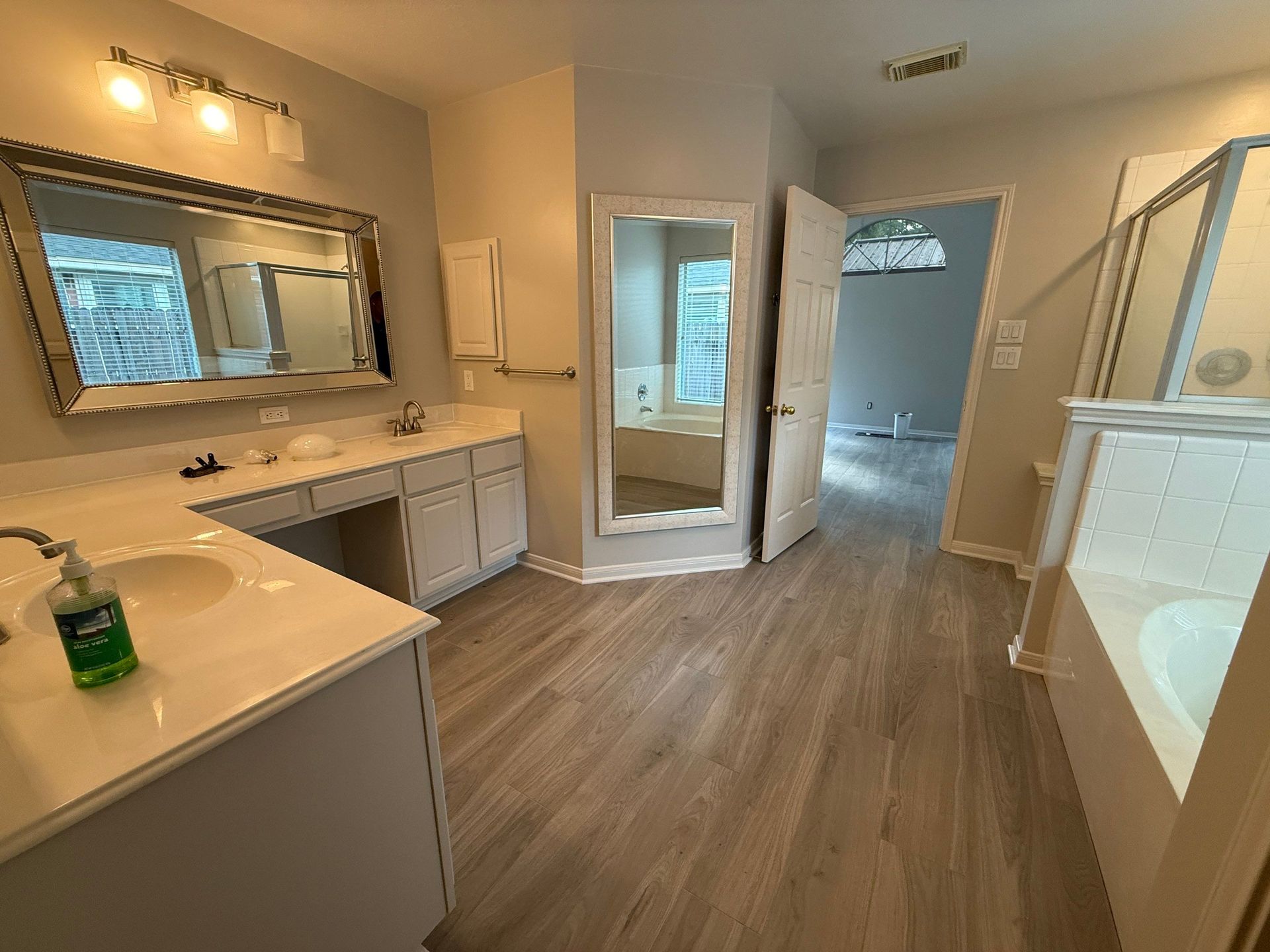 Bathroom with light wood-look flooring, dual sinks, large mirror, and open doorway to another room.