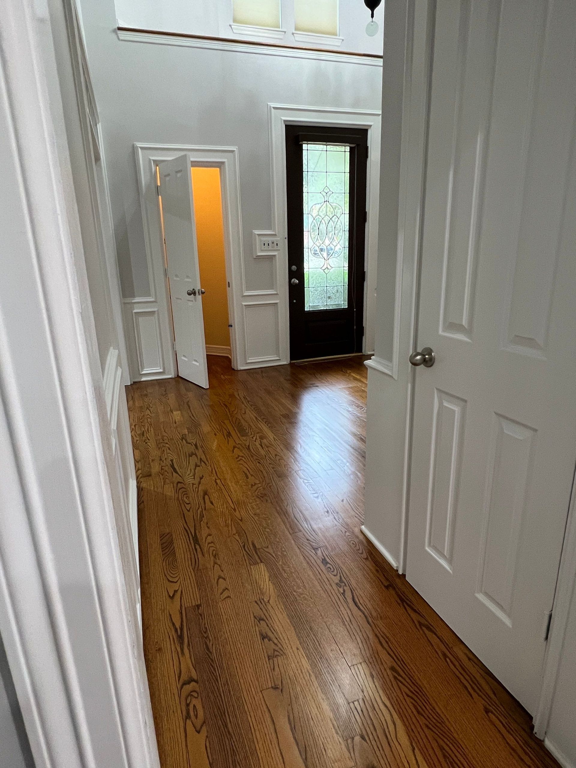 Hallway with hardwood floor, white trim, and a dark front door. A door is ajar, revealing a light-filled room.