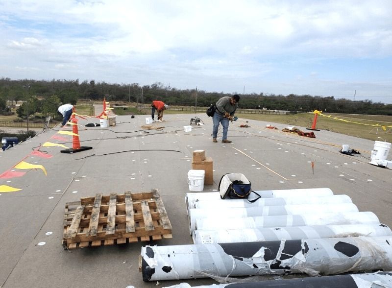 Workers on a flat roof, inspecting and working near rolled roofing materials, safety cones, and a wooden pallet under cloudy skies.