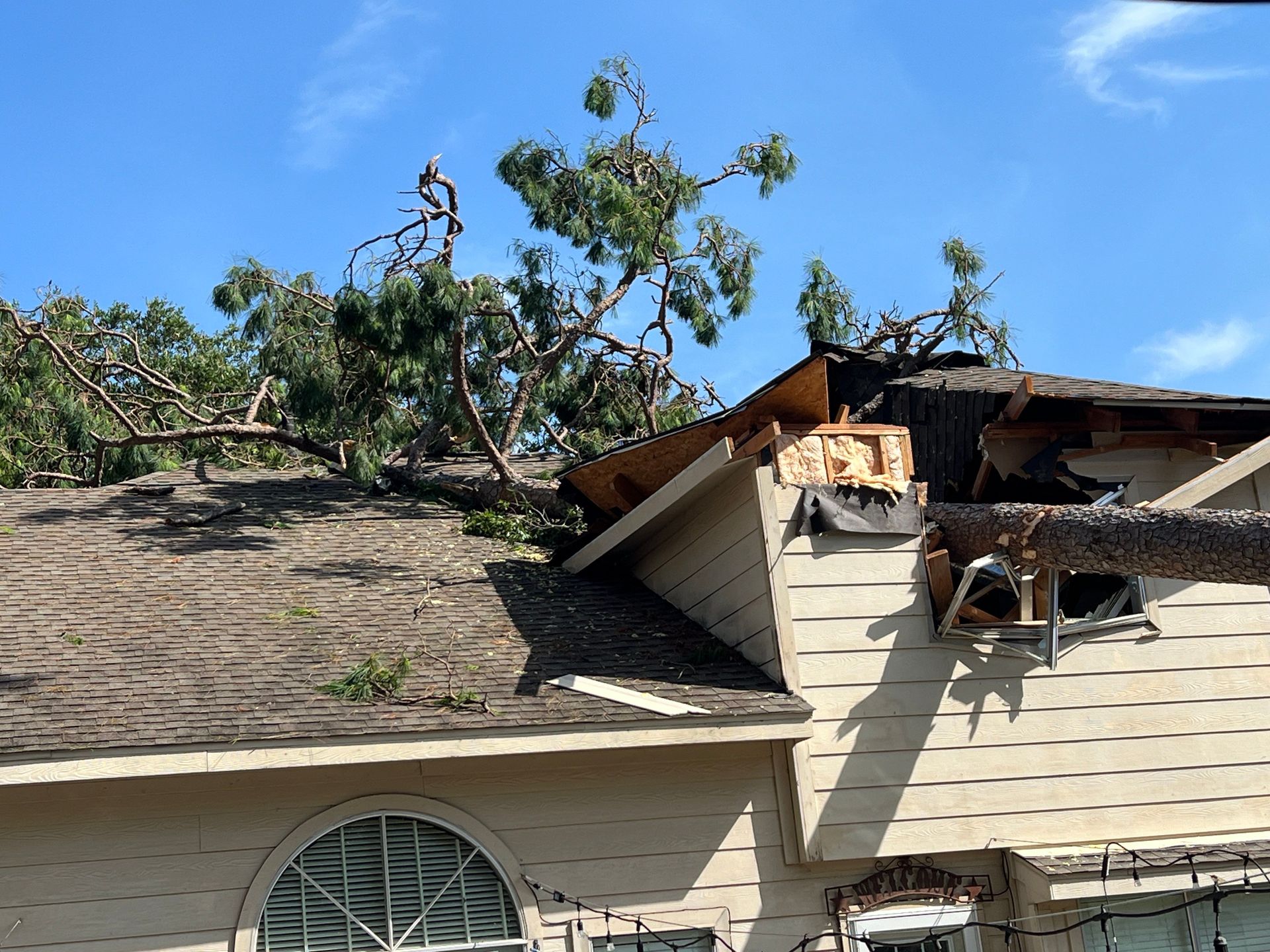 Tree branches on damaged roof of a house; brown roof, light siding, blue sky.