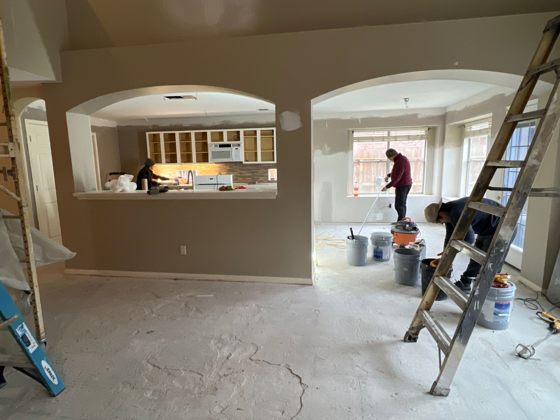 Interior view of a room under renovation, with workers, ladders, and paint buckets. Kitchen in the background.
