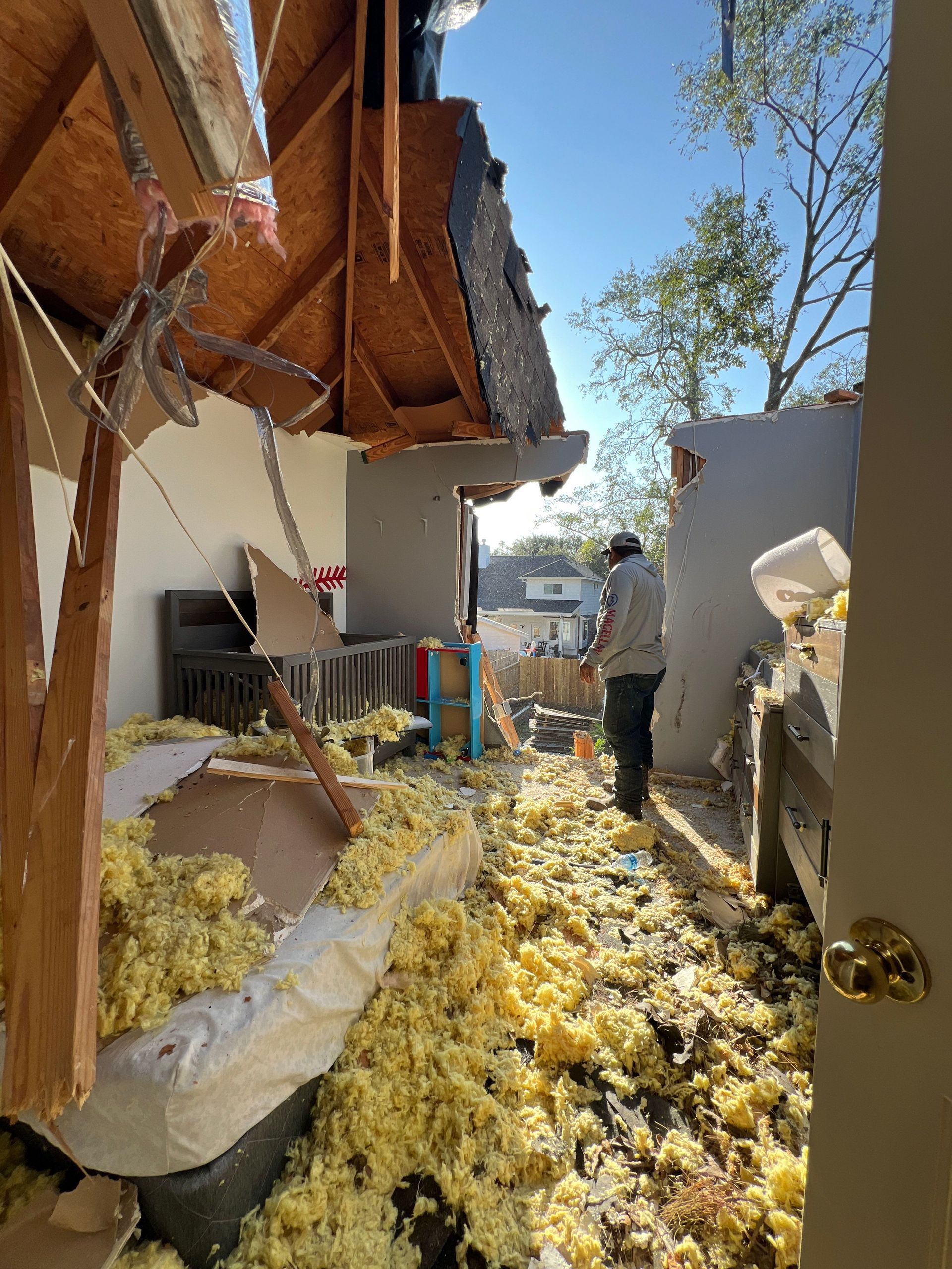 House damage after a storm: collapsed roof, debris, insulation strewn about, person surveying the damage.