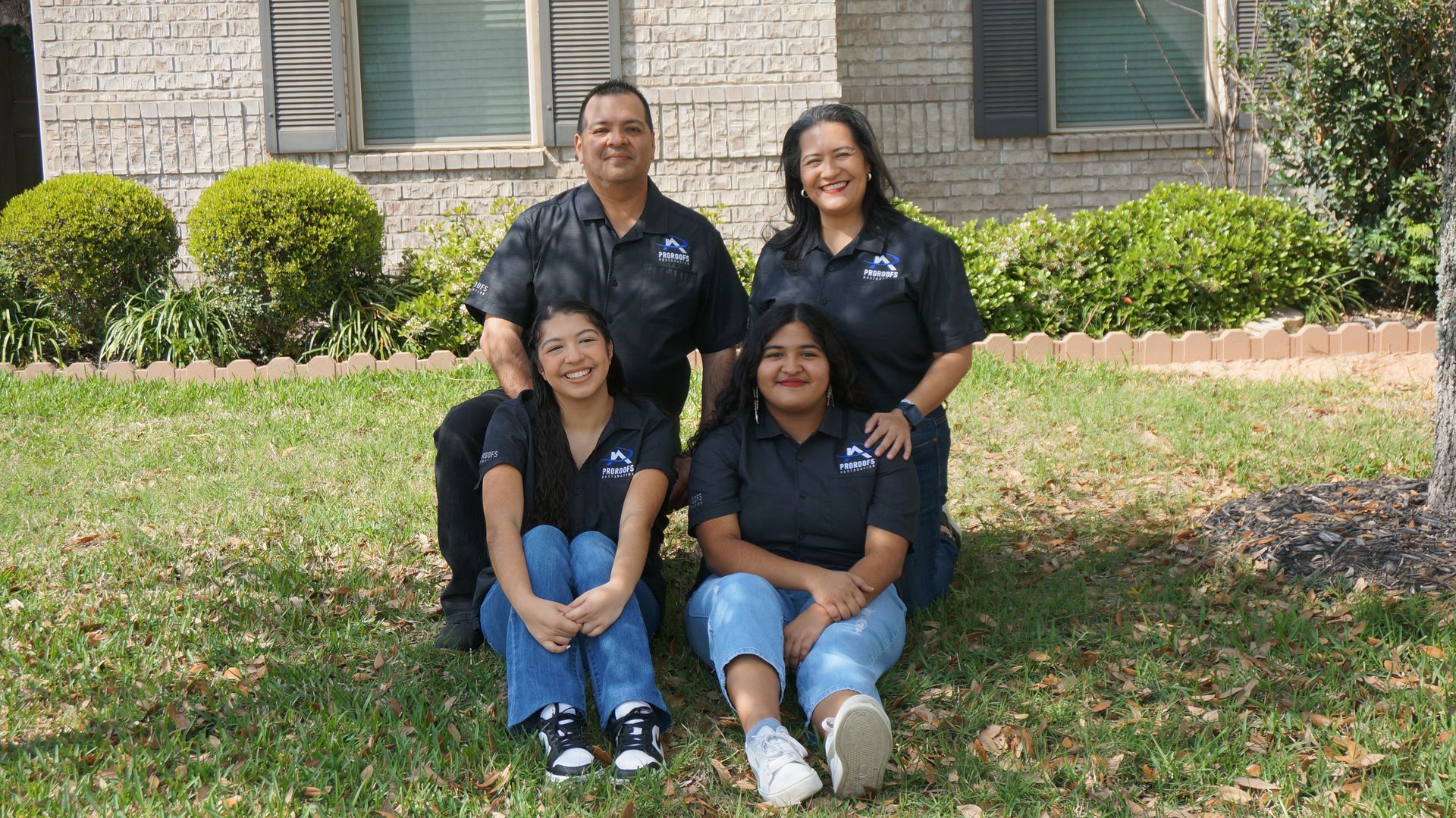 Family of four in front of a house, all wearing matching black shirts. Sitting on grass.