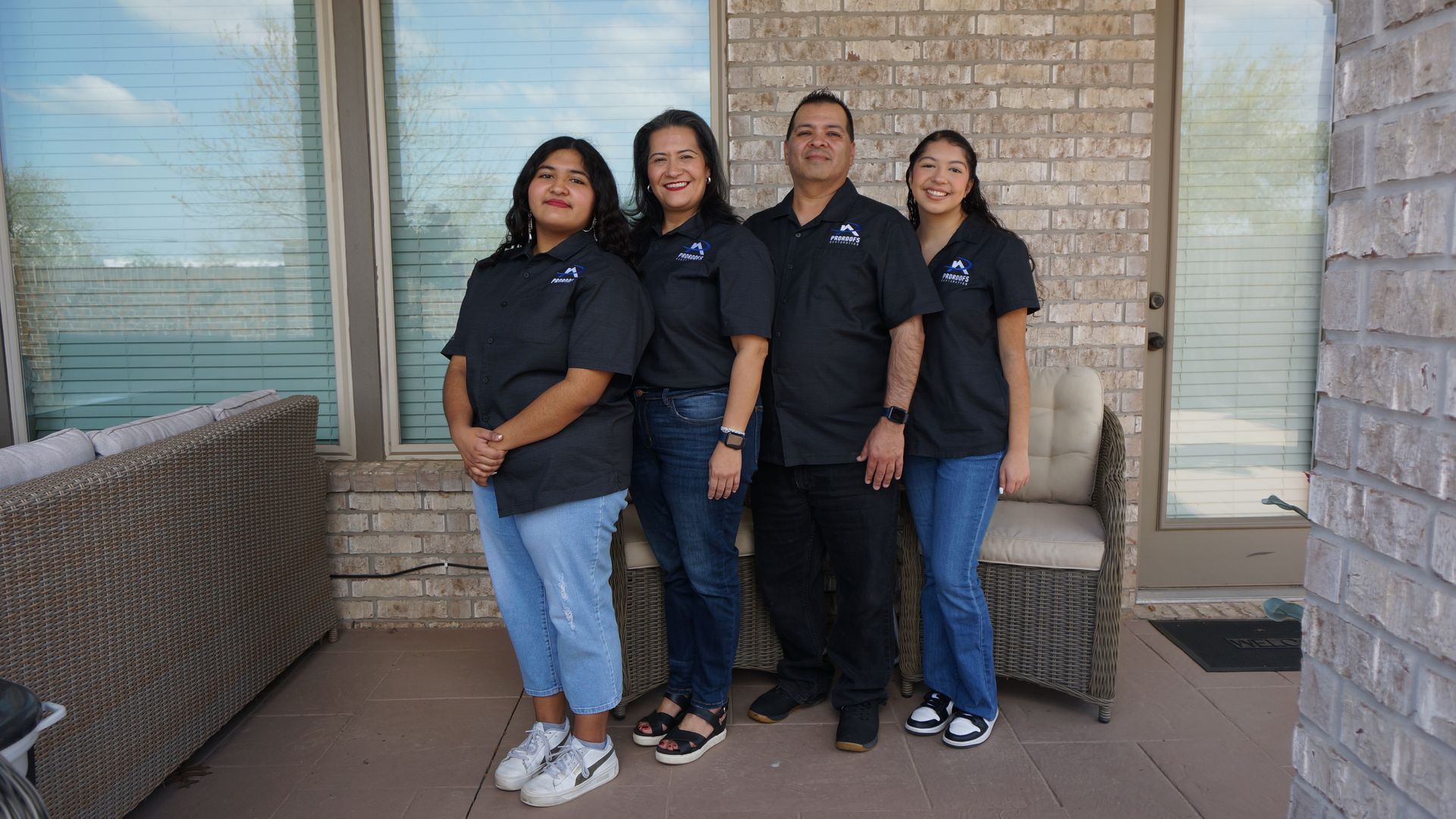 Family of four in matching black shirts pose outdoors near a brick wall.