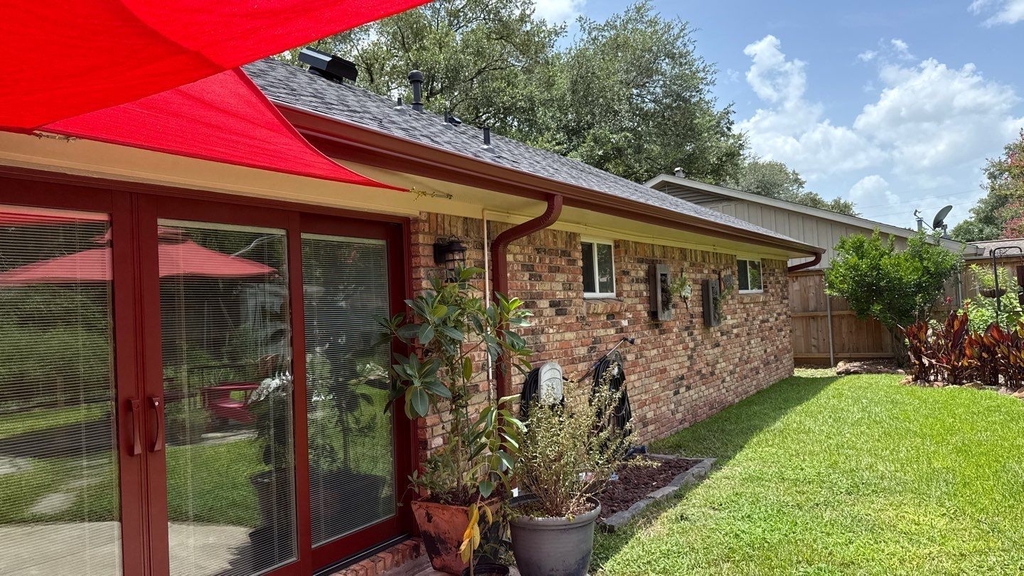 Brick house with red trim, gutters, and a red patio umbrella, with a green lawn and trees.