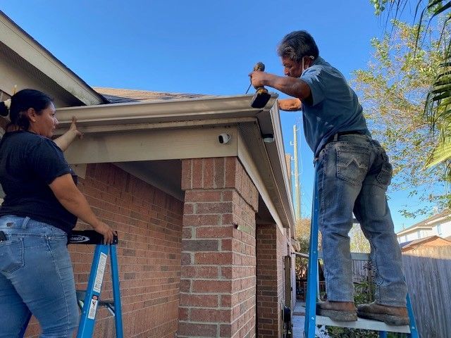 Two people installing gutters on a brick home; one on a ladder using a tool, the other holding the gutter.