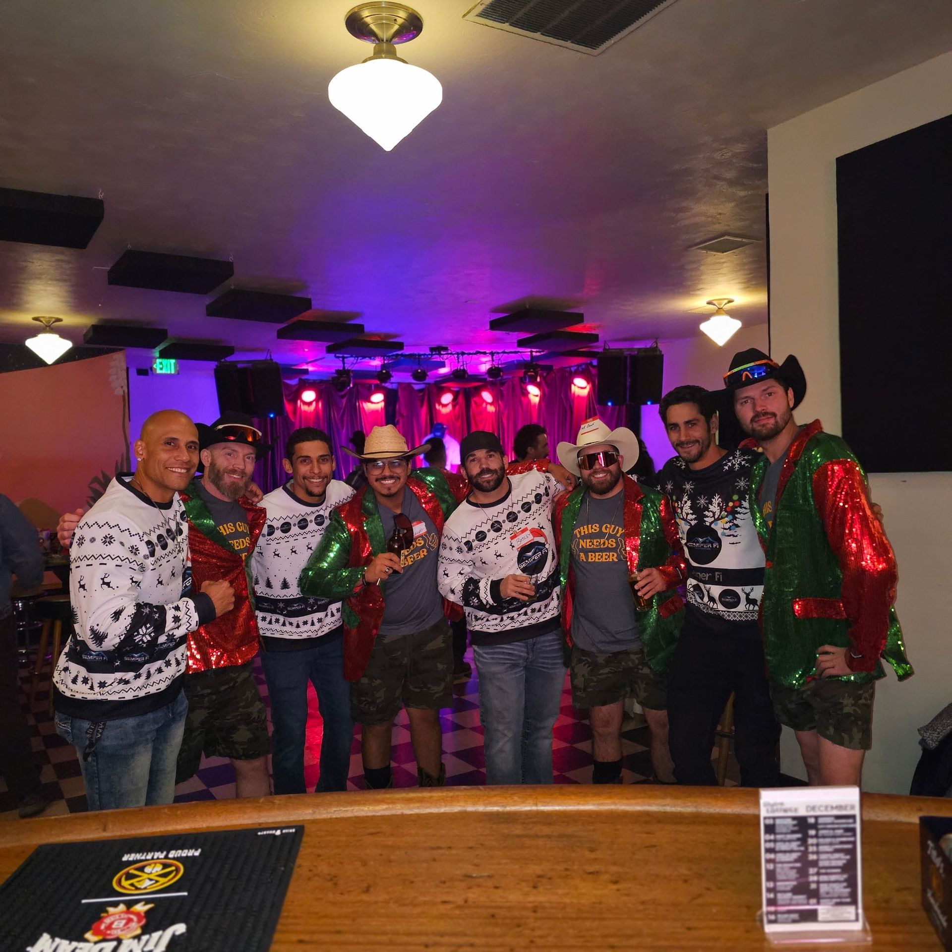 Group of people in Christmas sweaters at a bar, standing near a counter. Purple and blue stage lights in the background.