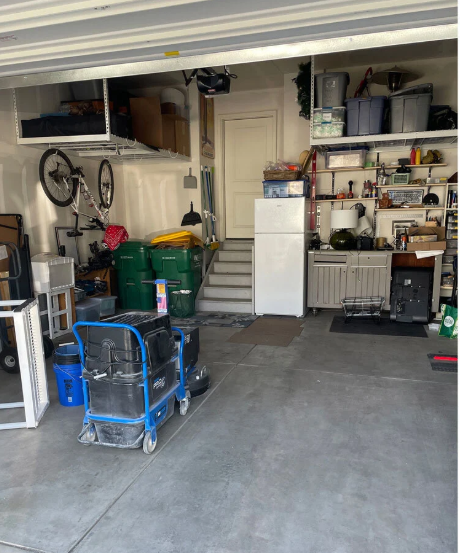 Cluttered garage with a cleaning machine, trash cans, shelving, and a refrigerator.