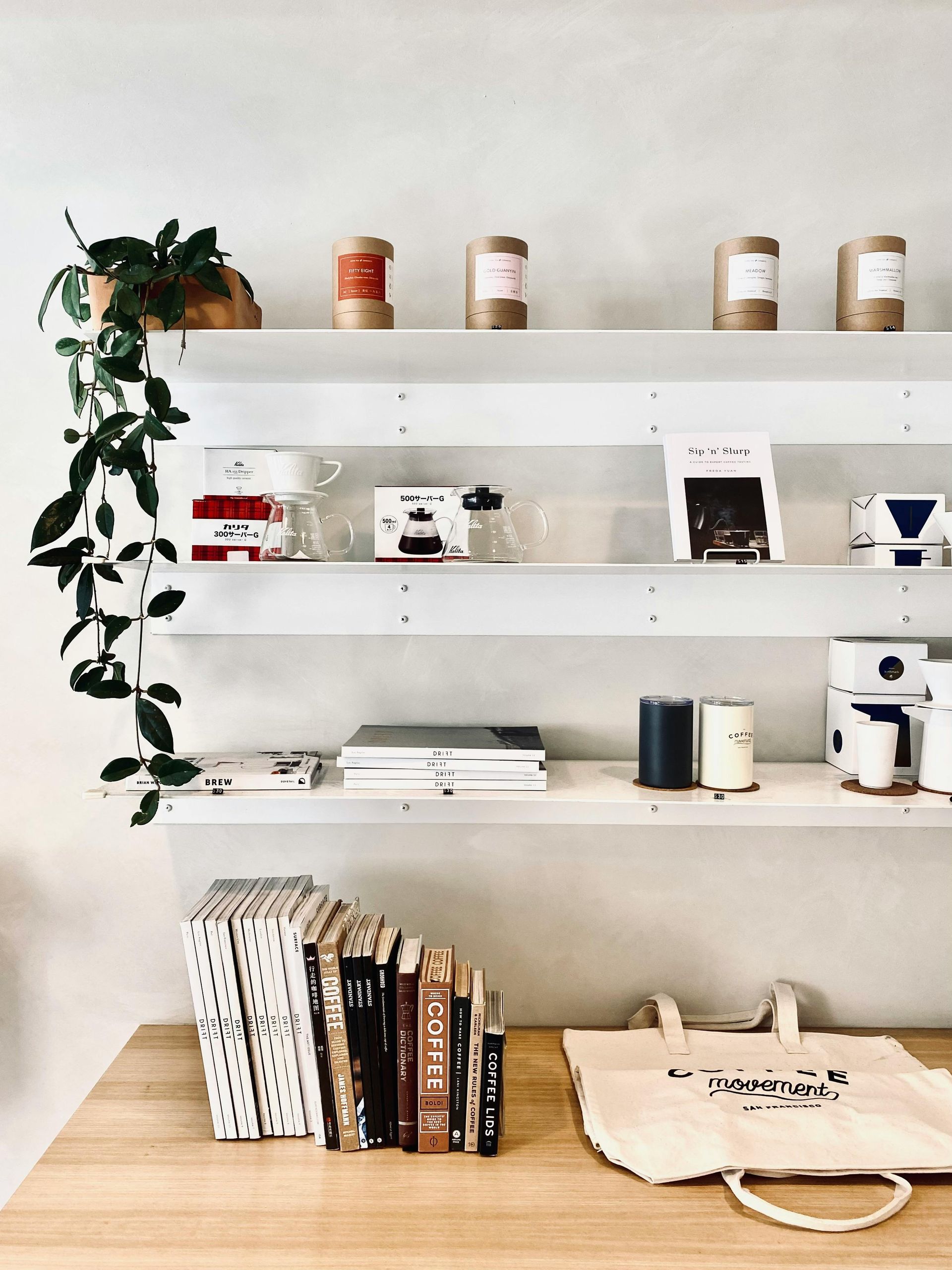 Shelves displaying coffee products and books in a shop. A trailing plant hangs, and a tote bag rests on a wooden table.