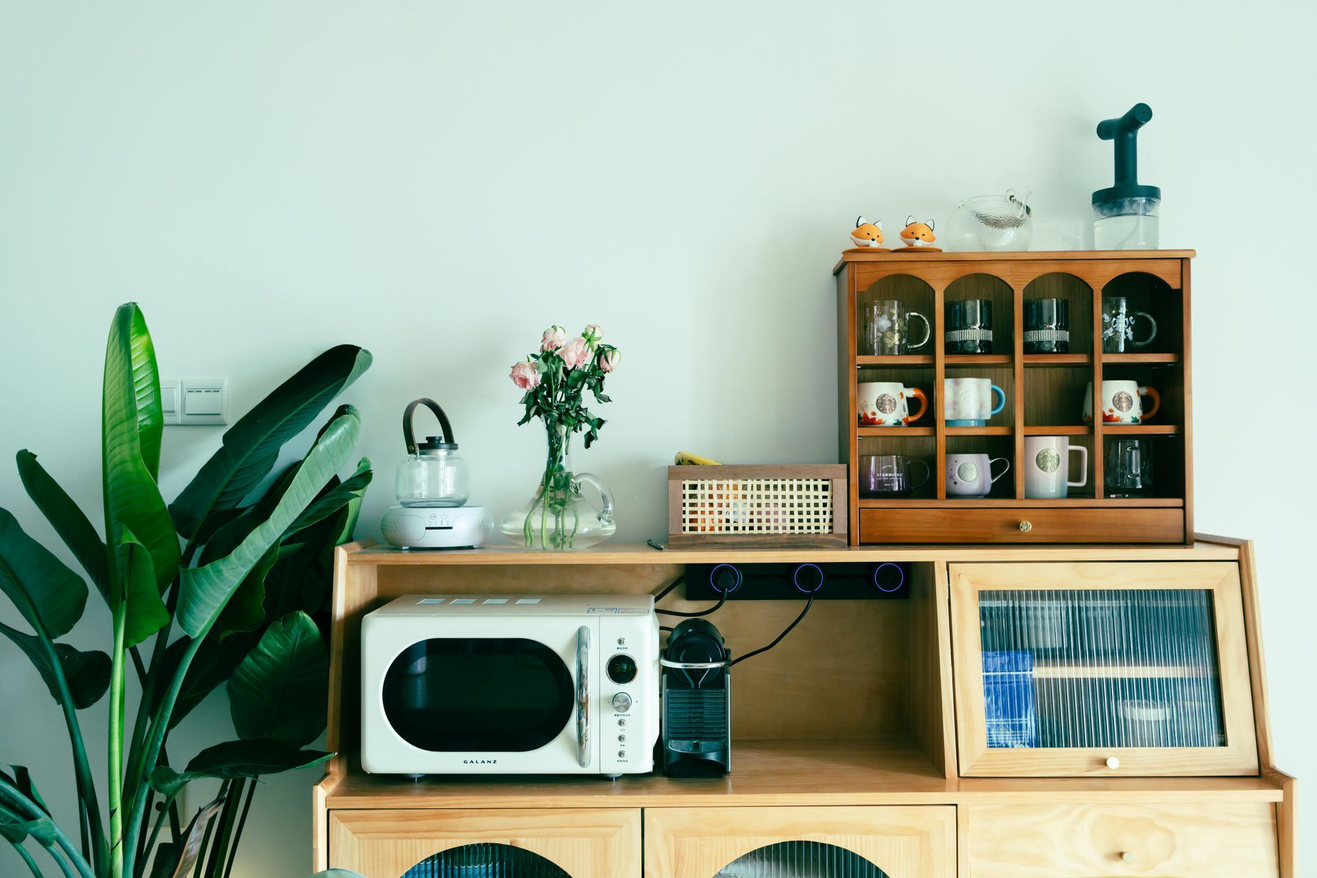 Wooden kitchen hutch with microwave, glassware, and plants against a white wall.