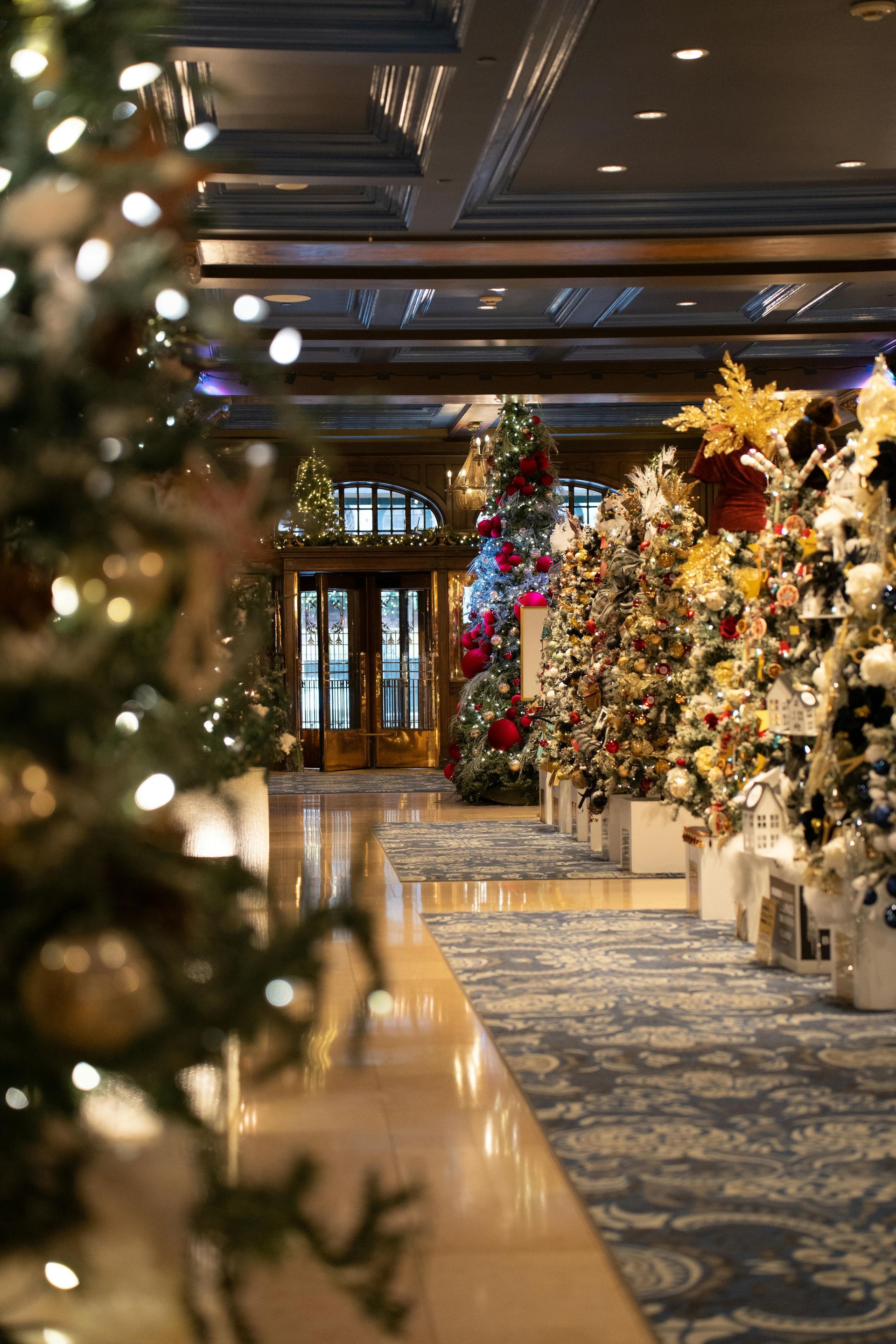 Hallway lined with decorated Christmas trees, leading to a door; gold and white lights sparkle.