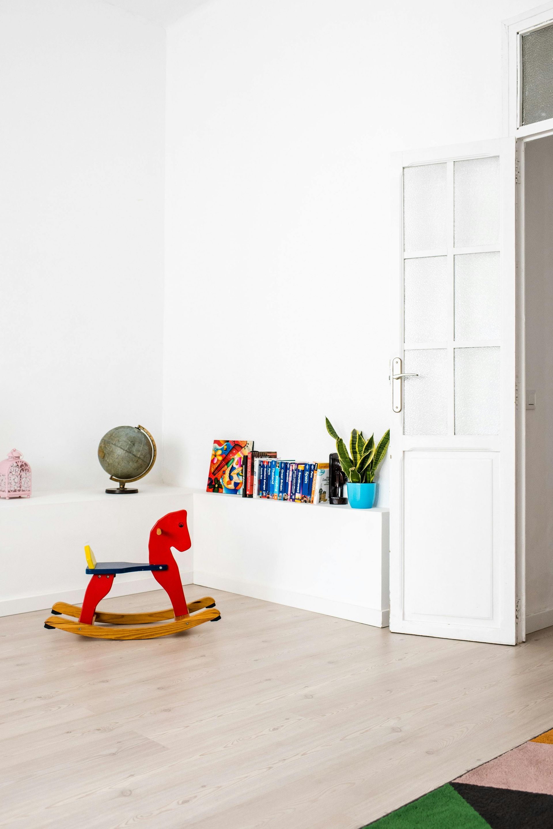 Red rocking horse in a white room with a shelf holding books, globe, and plant. Door visible.