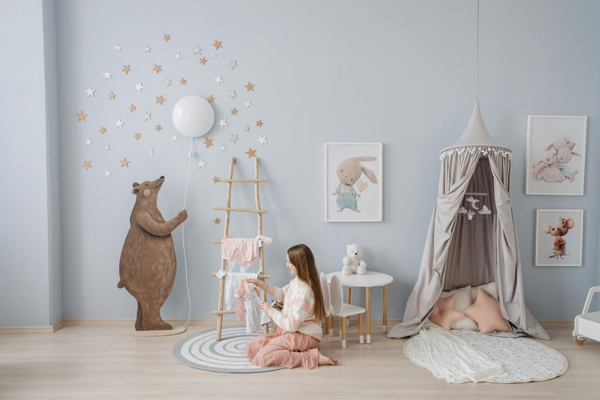 Girl in nursery arranges clothes on a ladder. Light blue walls, decorative bear, star decals, tent, artwork.