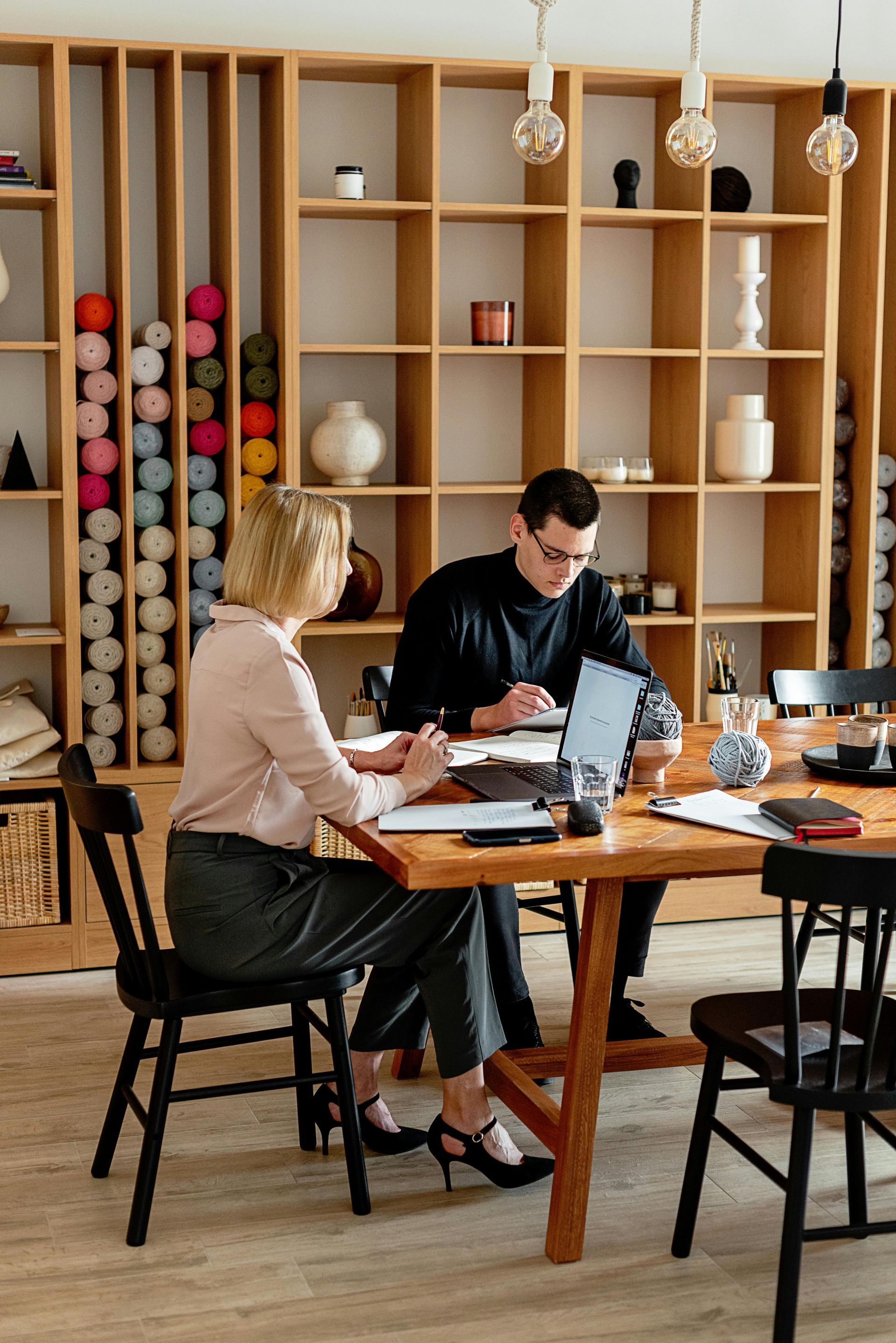 Two people working at a wooden table in a room with shelves. One person is using a laptop.