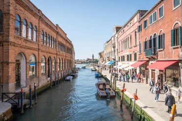 Un canal en Venecia con edificios de ladrillo rojo a ambos lados y gente paseando por la orilla bajo toldos rojos.