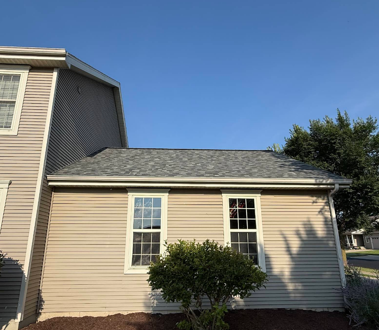 Tan-sided house with two windows and gray roof against a clear blue sky.