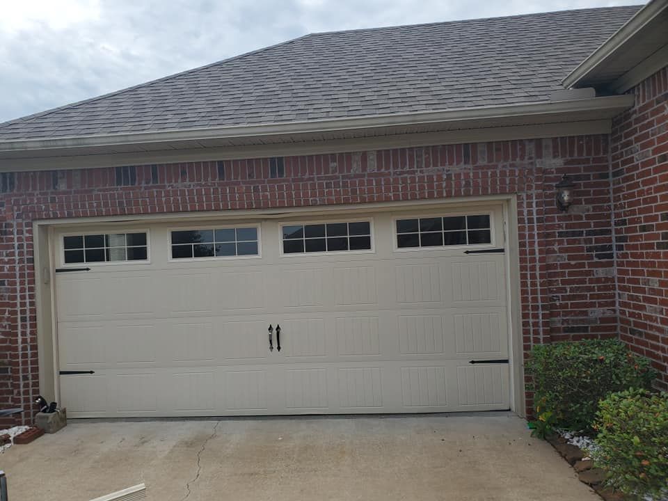 A beige garage door with decorative windows and black hardware on a brick home exterior.