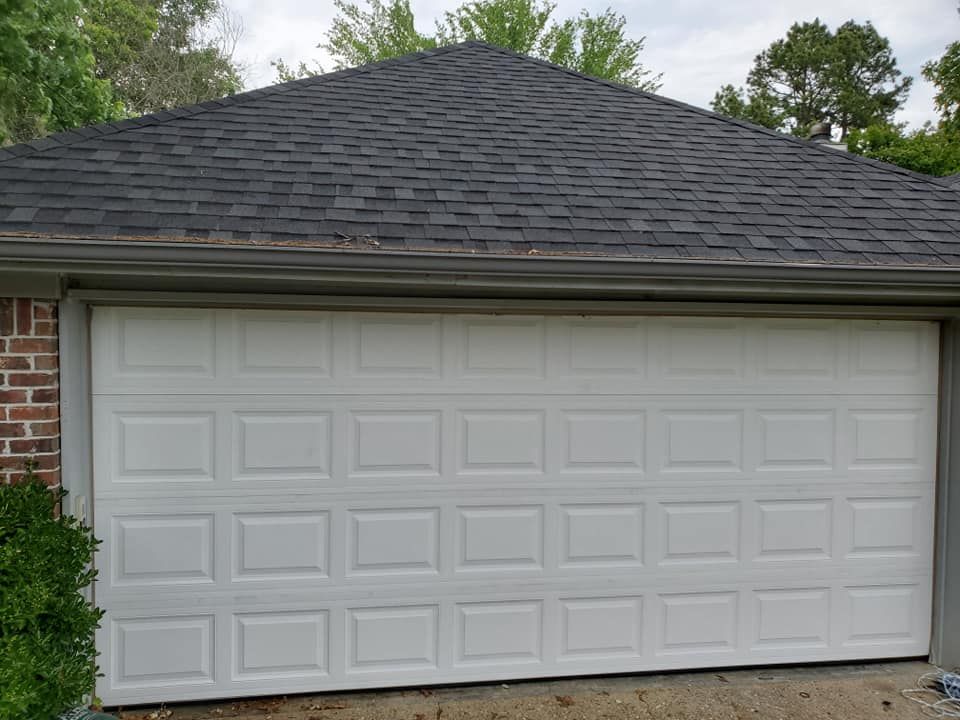 A white, multi-panel garage door installed under a dark-shingled roof and brick exterior wall.