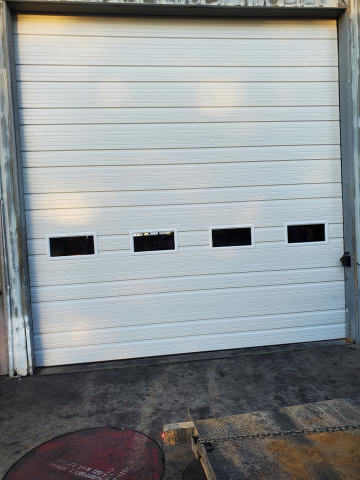 A closed white industrial overhead garage door with a row of four rectangular windows set in a concrete frame.