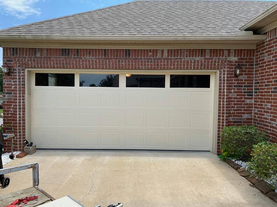 A white garage door with four rectangular windows at the top, set in a red brick exterior wall with a concrete driveway.