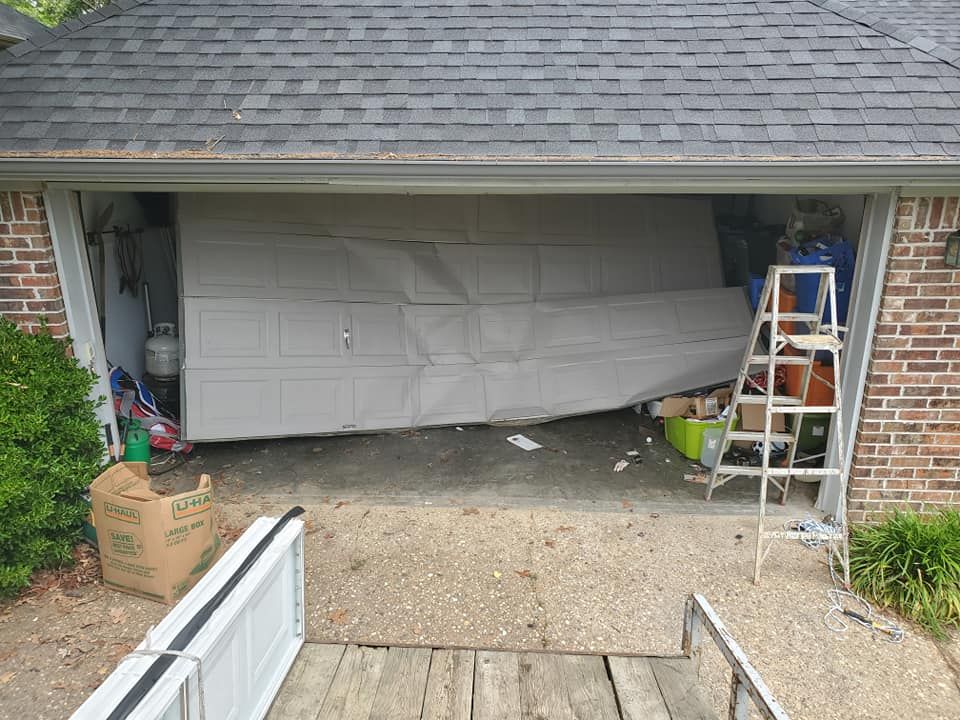 A damaged, buckled white garage door hangs unevenly in the frame of a brick house with a ladder nearby.